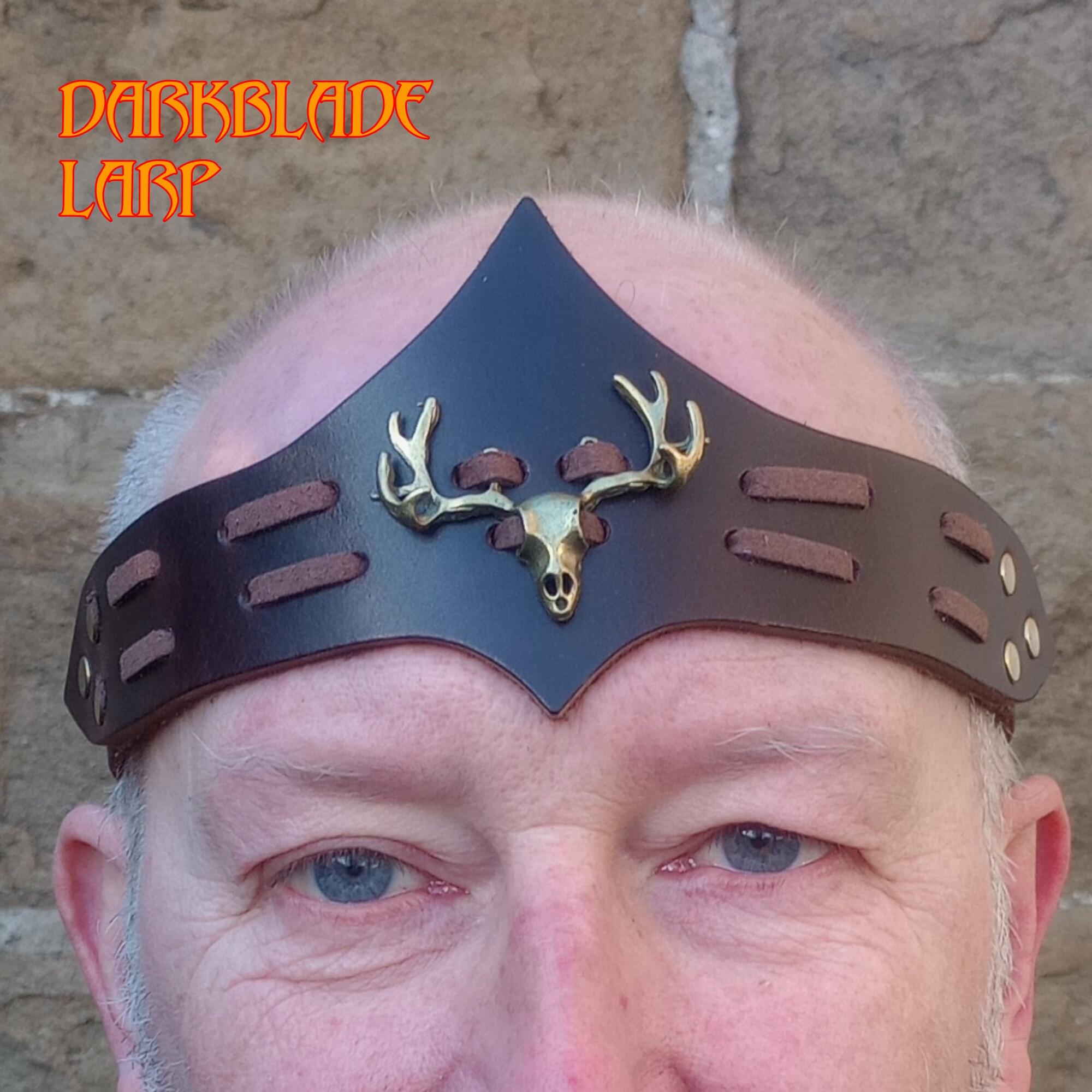 Face of a bearded man wearing a brown Leather headband with a brass stag skull with antlers laced to it. The headbandpeaks to a point top and bottom on the centreline.