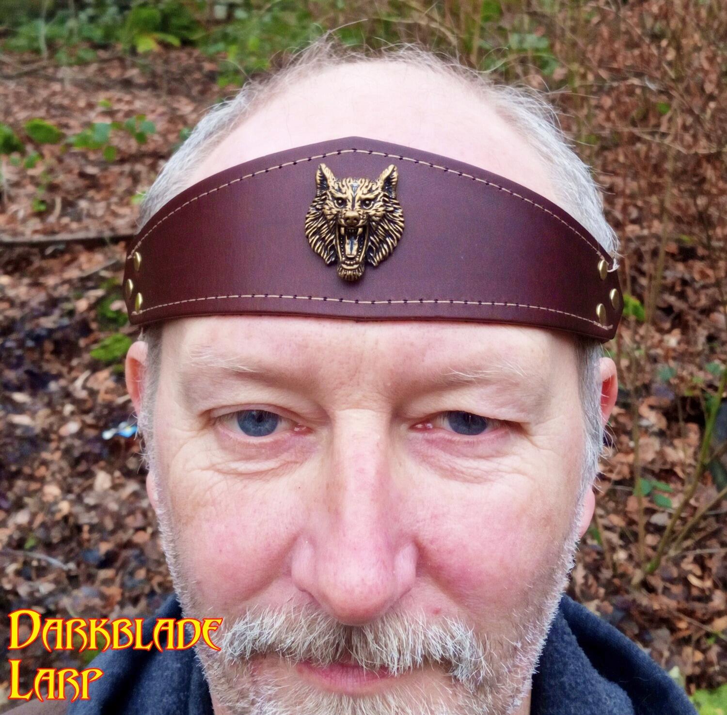 Face of a bearded man wearing a brown Leather headband with a brass snarling wolf head on it. There is pale stitching round the edge of the leather. The headband tapers out high point on the top on the centreline.