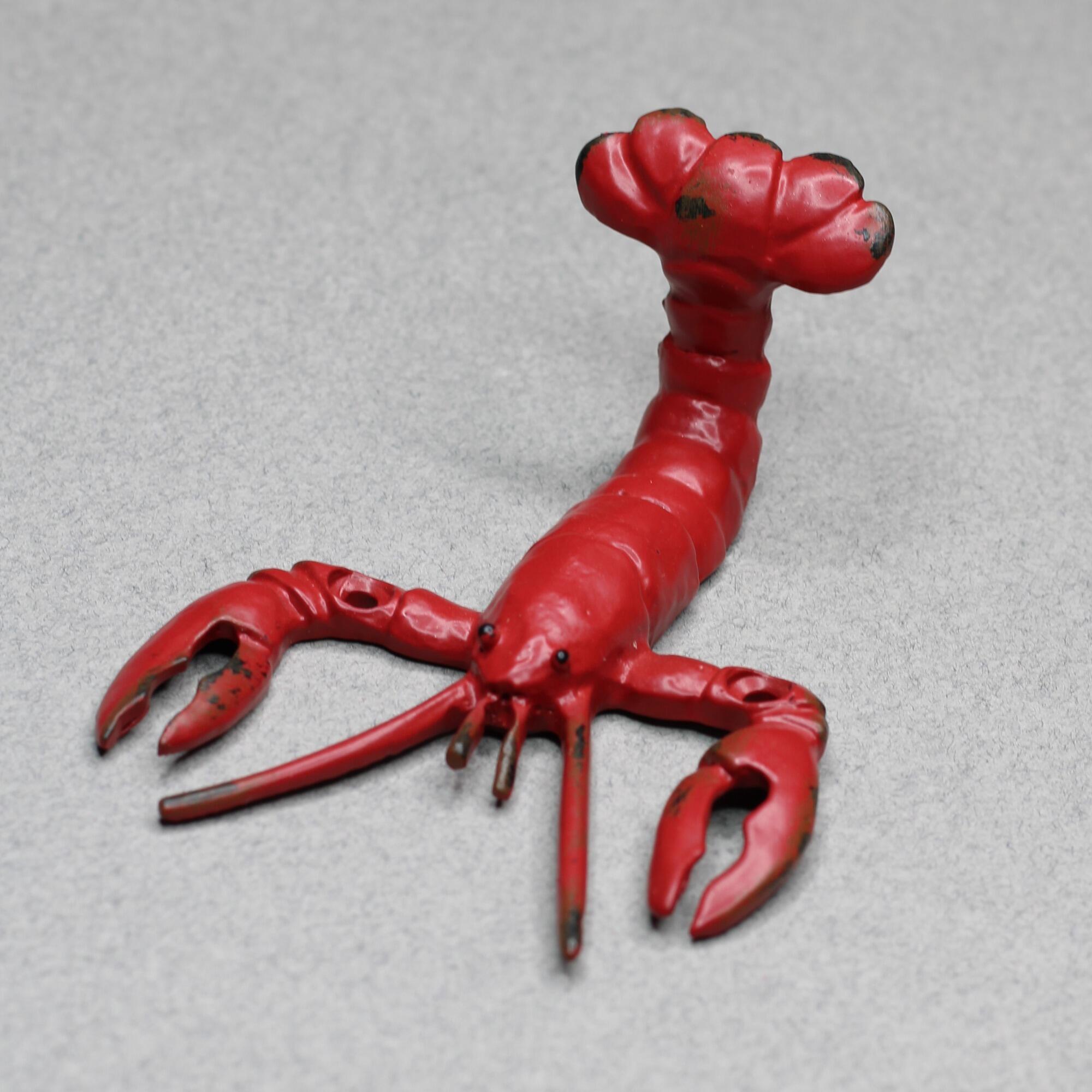 A red lobster-shaped coat hook lying on a flat surface, highlighting the three-dimensional curved tail design and rustic black distressed paint accents.