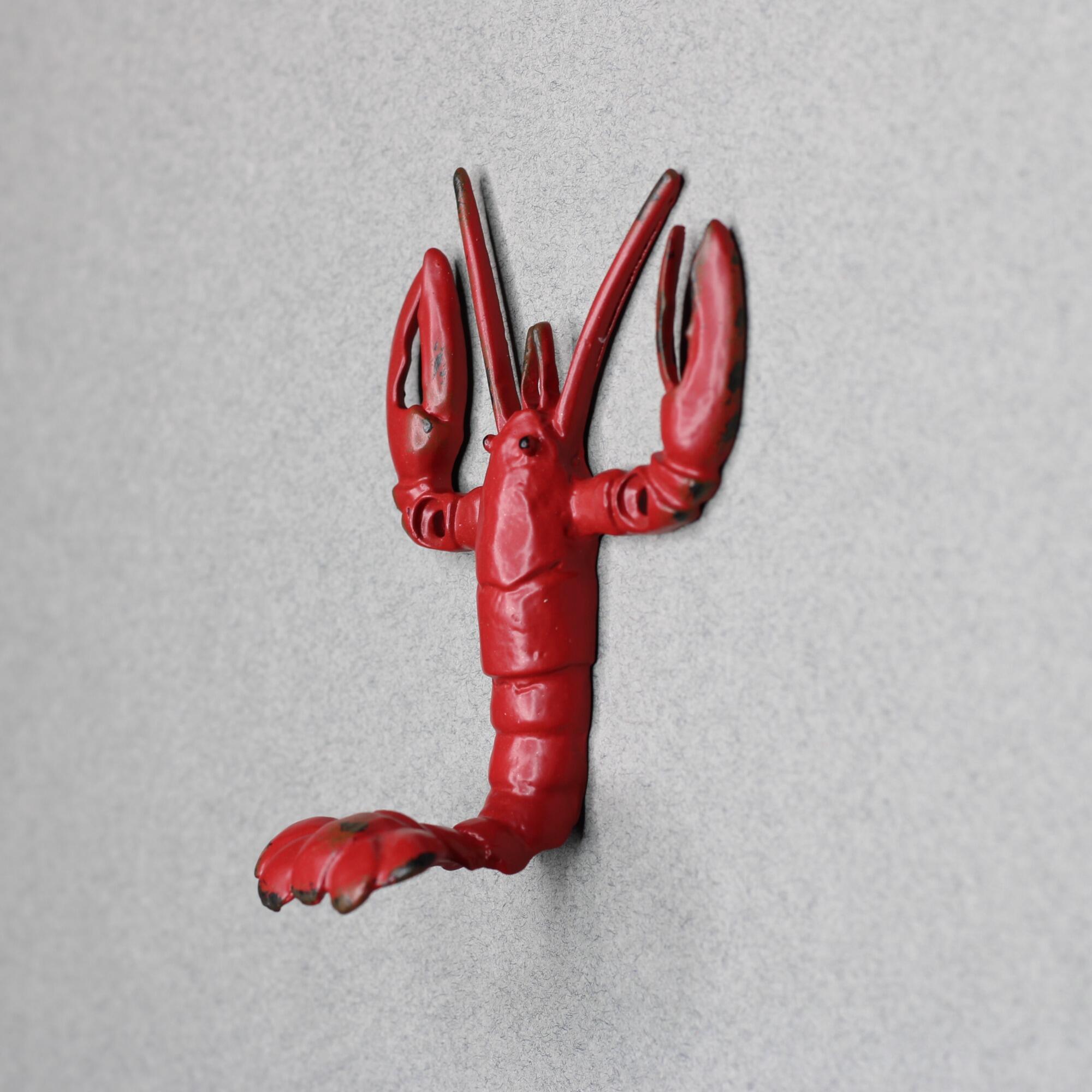 Side view of a vintage-style red metal lobster wall hook mounted on a gray wall, showing the lobster's tail curving outward to form a sturdy hanging hook.