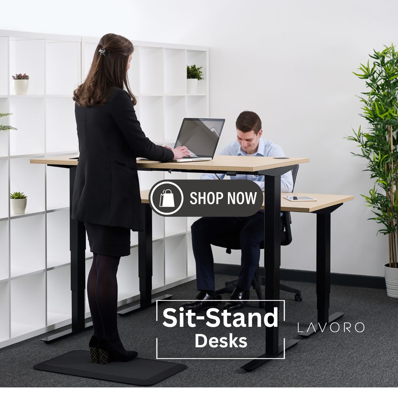 Modern open-plan office space with people working at height adjustable desks in seated and standing positions, featuring large windows and ergonomic furniture.