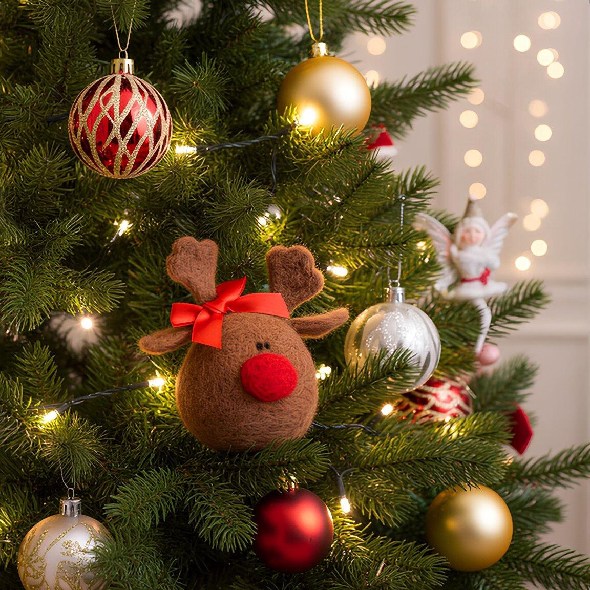 A round, brown needle-felted reindeer named Rosie with a large red nose, small antlers, and a bright red satin bow, featured as a Christmas tree decoration surrounded by festive lights and baubles.