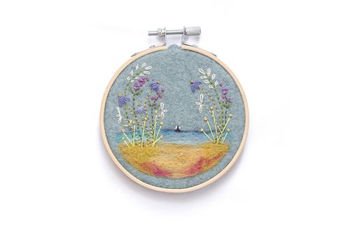 Completed seaside embroidery in a wooden hoop featuring a needle-felted beach and ocean background on blue felt with embroidered coastal flowers.