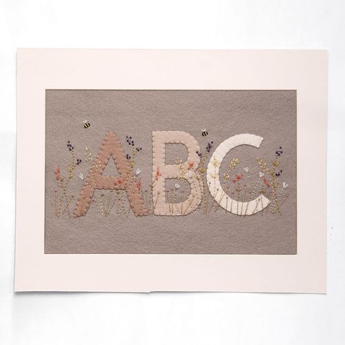 Completed embroidery project featuring felt letters A, B, and C in shades of mushroom and stone grey, layered onto a beige felt background with embroidered purple wildflowers and bees