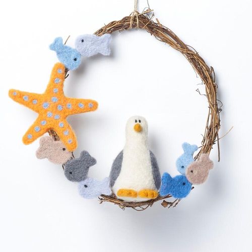 Nautical-themed needle felting kit featuring a round twig base, a white and grey 3D seagull with orange feet, a spotted orange starfish, and a school of blue, grey, and sand-colored felted fish.