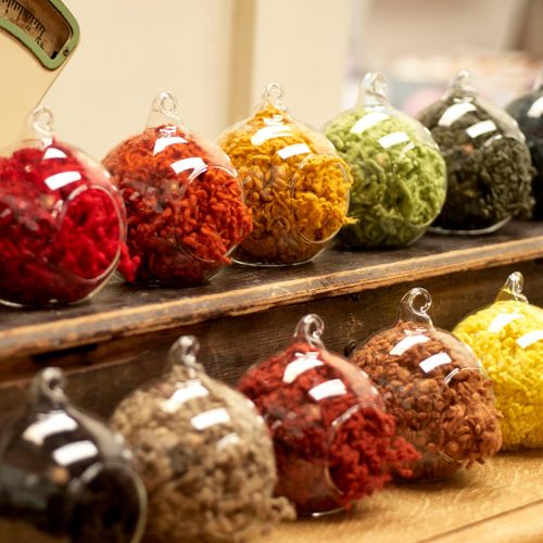 A row of glass apothecary jars filled with colourful wool nepps in shades of red, mustard, green, and brown displayed on a rustic wooden shelf.