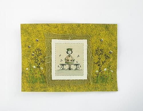 Completed embroidery project featuring a whimsical illustration of a little girl sitting on a garden bench, layered onto yellow mustard felt with embroidered wildflowers and bees.