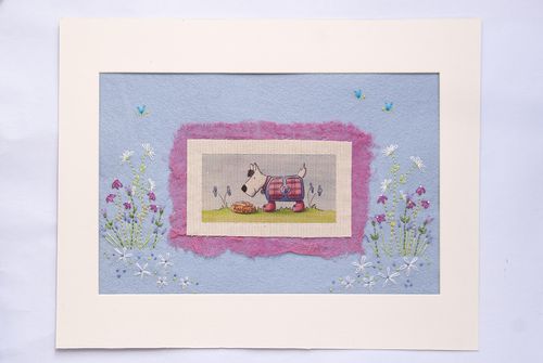 Completed embroidery project featuring an illustration of a white terrier dog named Angus in a tartan coat, layered onto light blue felt with pink textured fabric and embroidered white and purple flowers.