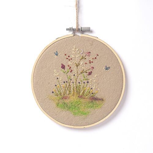 Completed floral embroidery in a wooden hoop featuring a natural beige felt background with a needle-felted meadow base, red blossoms, and blue dragonflies.