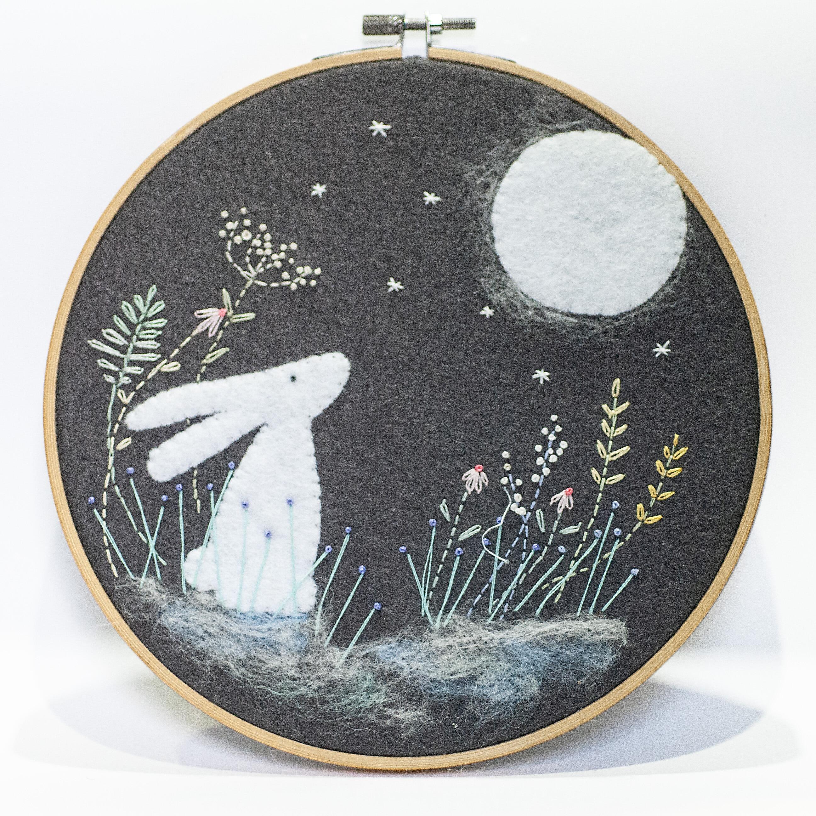 Completed embroidery project in a wooden hoop featuring a hand-cut felt white hare and full moon on a charcoal grey felt background with embroidered stars and meadow flowers.