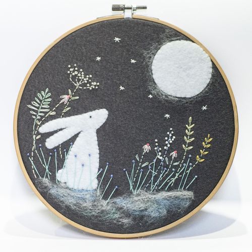 Completed embroidery project in a wooden hoop featuring a hand-cut felt white hare and full moon on a charcoal grey felt background with embroidered stars and meadow flowers.