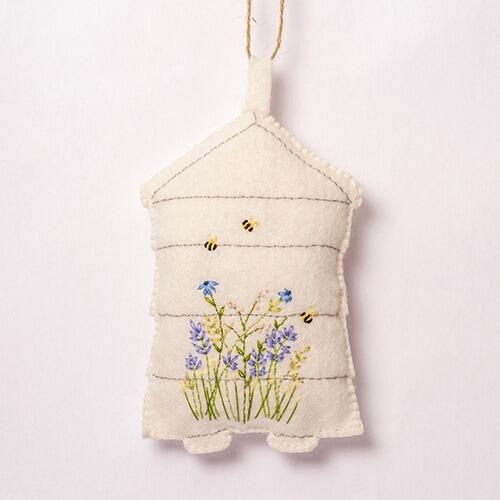 A 3D padded cream felt beehive featuring horizontal stitched lines, embroidered purple lavender and blue wildflowers, and three small embroidered