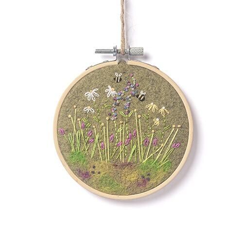 Completed floral embroidery in a wooden hoop featuring a lush green felt background with needle-felted grass, purple foxgloves, white daisies, and bees.
