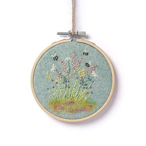 Completed floral embroidery in a wooden hoop featuring a soft needle-felted meadow base on aqua blue felt with embroidered daisies, pink blossoms, and bees.