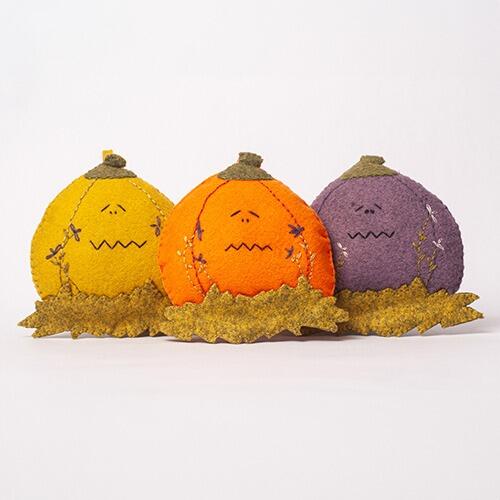 Three 3D padded felt pumpkins named Eerie, Creepy, and Boo in yellow, orange, and purple, each featuring unique embroidered faces and sitting on a bed of felt autumn grass.