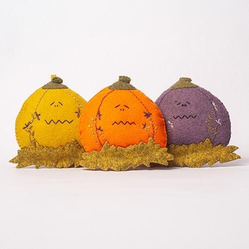 Three 3D padded felt pumpkins named Eerie, Creepy, and Boo in yellow, orange, and purple, each featuring unique embroidered faces and sitting on a bed of felt autumn grass.
