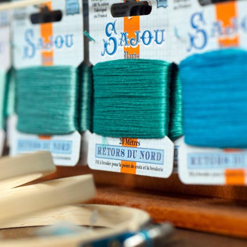 Close-up of Maison Sajou Retors du Nord embroidery thread cards in teal, turquoise, and aqua blue on a wooden display.