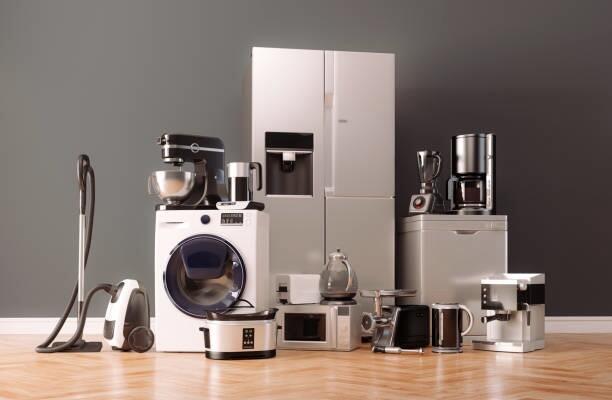 Assorted kitchen appliances including a fridge and washing machine displayed on a wooden floor against a grey wall