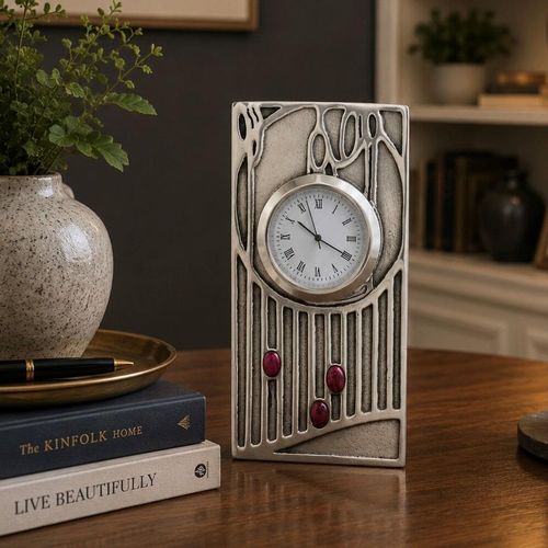 An Art Nouveau style pewter desk clock influenced by Charles Rennie Mackintosh, featuring a tall rectangular frame with a textured finish and stylized vertical line work. The circular white clock face with Roman numerals is set into the upper half, while the base is accented with three vertical rows ending in small, purple cabochon gems
