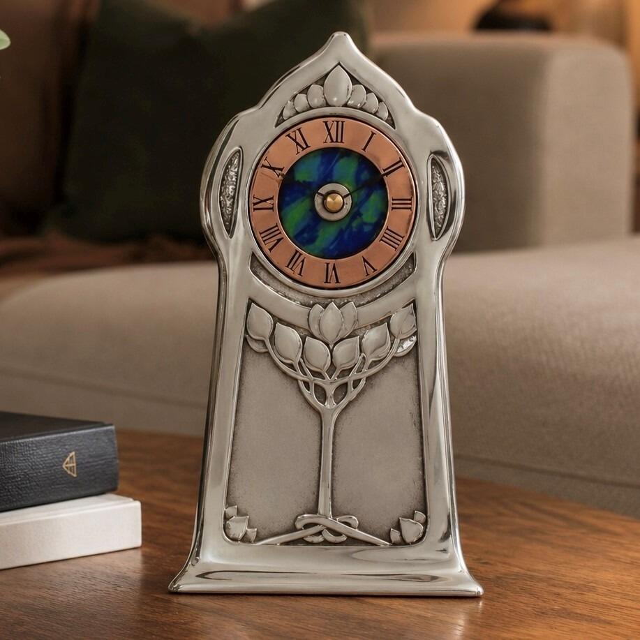An Arts and Crafts style pewter mantel clock designed by Archibald Knox, featuring a copper chapter ring with Roman numerals surrounding a vibrant blue and green enamelled centre. The polished pewter case has a tall, arched silhouette decorated with an embossed stylized tree design