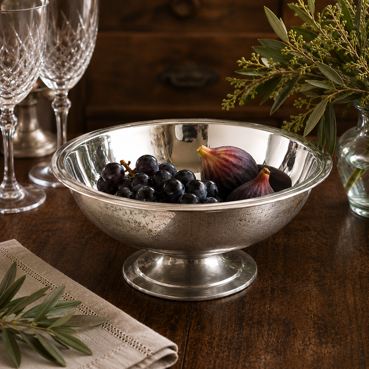 Handcrafted pewter footed bowl with a bright mirror-polished interior and lightly antique-textured exterior, displayed on a wooden dining table filled with dark grapes and fresh figs, surrounded by crystal stemware and greenery.