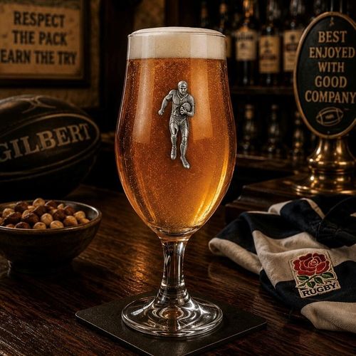 A stemmed beer chalice glass filled with golden ale and a frothy white head, featuring a raised pewter emblem of a rugby player running with a ball
