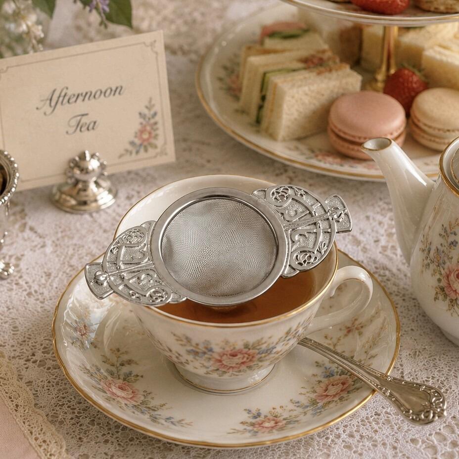 A stainless steel mesh tea strainer with ornate pewter handles featuring an Art Nouveau Celtic knot design, resting across a floral porcelain teacup during an afternoon tea service