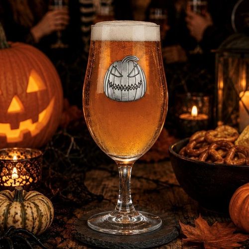 A stemmed beer glass filled with amber ale and a thick foam head, featuring a detailed pewter evil pumpkin jack-o'-lantern emblem on the front