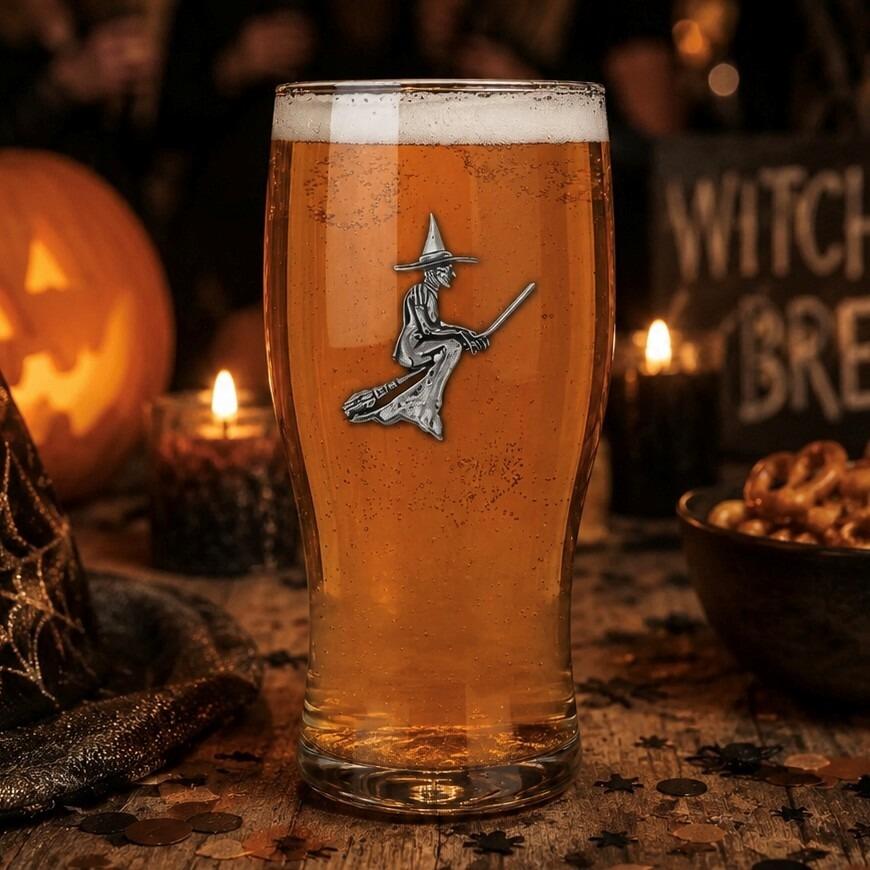 A pint glass filled with amber beer and a white foam head, featuring a detailed pewter emblem of a witch flying on a broomstick