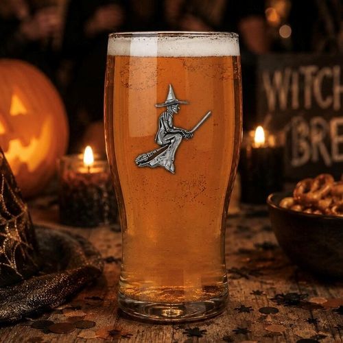A pint glass filled with amber beer and a white foam head, featuring a detailed pewter emblem of a witch flying on a broomstick