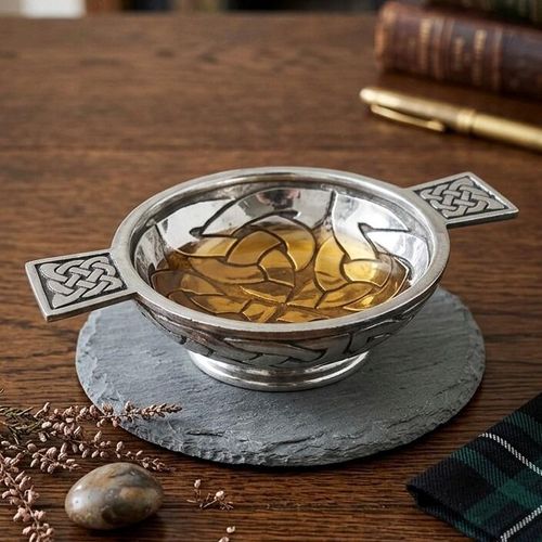 Traditional pewter quaich bowl filled with whiskey, featuring rectangular handles and an interior base decorated with Celtic knotwork patterns, displayed on a round slate mat with a tartan cloth