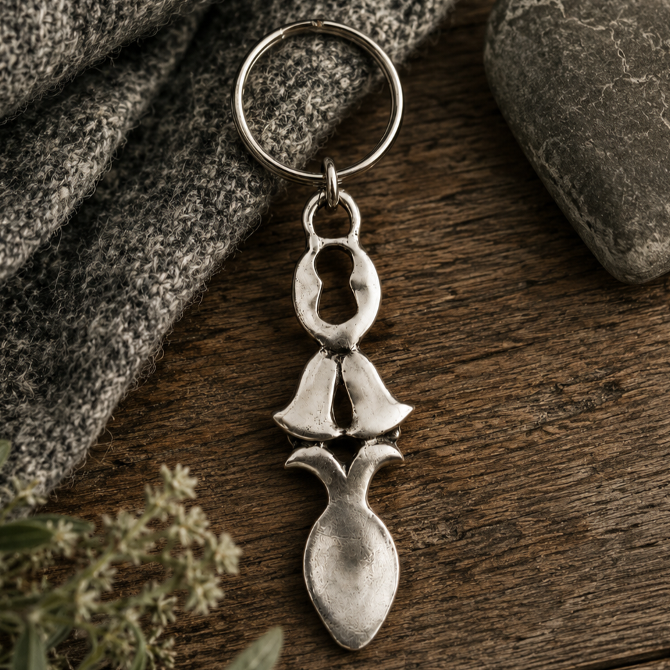 Handcrafted pewter Welsh lovespoon keyring featuring a keyhole cut-out, wedding bells, and a spoon bowl, resting on a rustic wooden background next to grey knit fabric and a smooth stone.