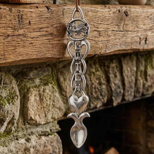 Handcrafted pewter Welsh lovespoon hanging against a wooden beam and stone hearth, featuring a circular Welsh dragon top, the word "CWTCH" integrated into open Celtic knotwork, a smooth solid heart, and a spoon bowl.