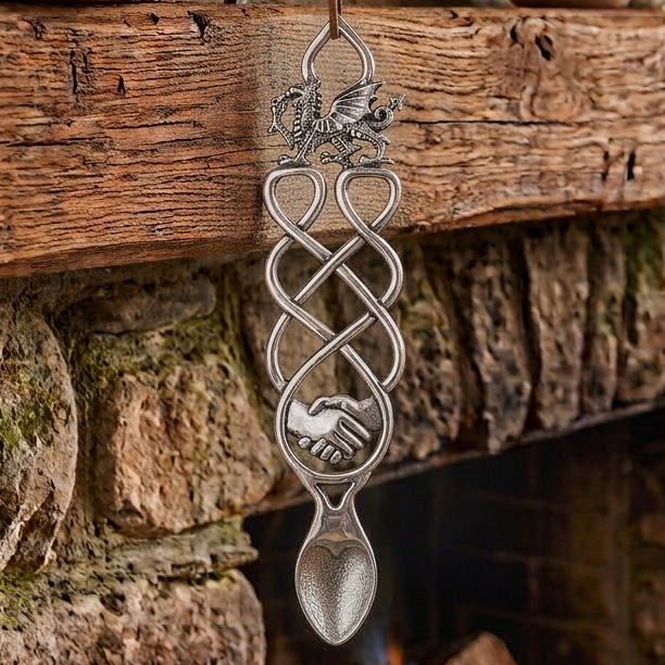 Handcrafted pewter Welsh lovespoon hanging against a wooden beam and stone fireplace, featuring a detailed Welsh dragon top, flowing open Celtic knotwork, a circular motif enclosing handshaking hands, and a textured spoon bowl.