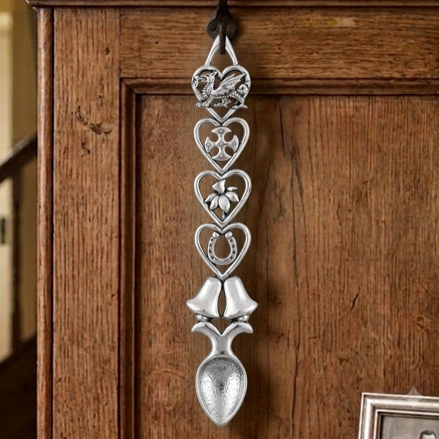 Handcrafted pewter Welsh lovespoon hanging on a wrought iron hook against a wooden panelled wall, featuring heart-shaped frames enclosing a Welsh dragon, a Celtic cross, a daffodil, a lucky horseshoe, and twin wedding bells above a textured spoon bowl.