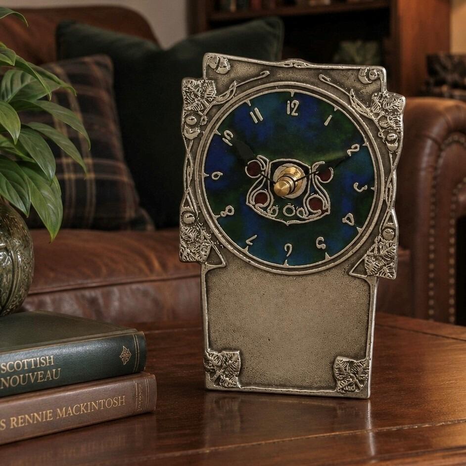 An Arts and Crafts style pewter mantel clock designed by Archibald Knox, featuring a vibrant blue and green enamelled circular face with stylized numerals and a Celtic-inspired centre motif. The rectangular pewter frame is decorated with embossed leafy corner details