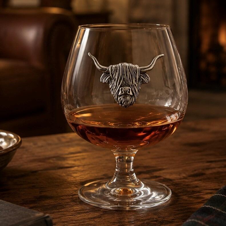 A short-stemmed clear glass brandy snifter filled with amber brandy, resting on a rustic wooden table. The front of the glass bowl is adorned with a detailed pewter relief emblem of a shaggy Highland cow's head with long, upward-curving horns