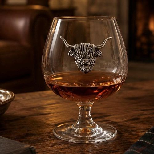 A short-stemmed clear glass brandy snifter filled with amber brandy, resting on a rustic wooden table. The front of the glass bowl is adorned with a detailed pewter relief emblem of a shaggy Highland cow's head with long, upward-curving horns