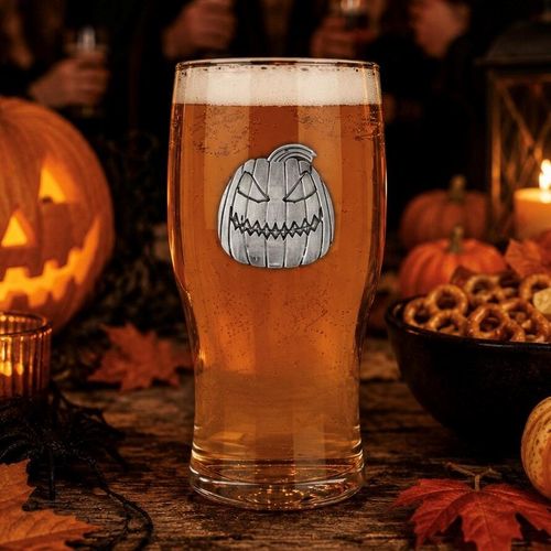 A clear straight pint beer glass filled with golden amber ale and a thick white head, featuring a detailed pewter jack-o'-lantern pumpkin motif in the center