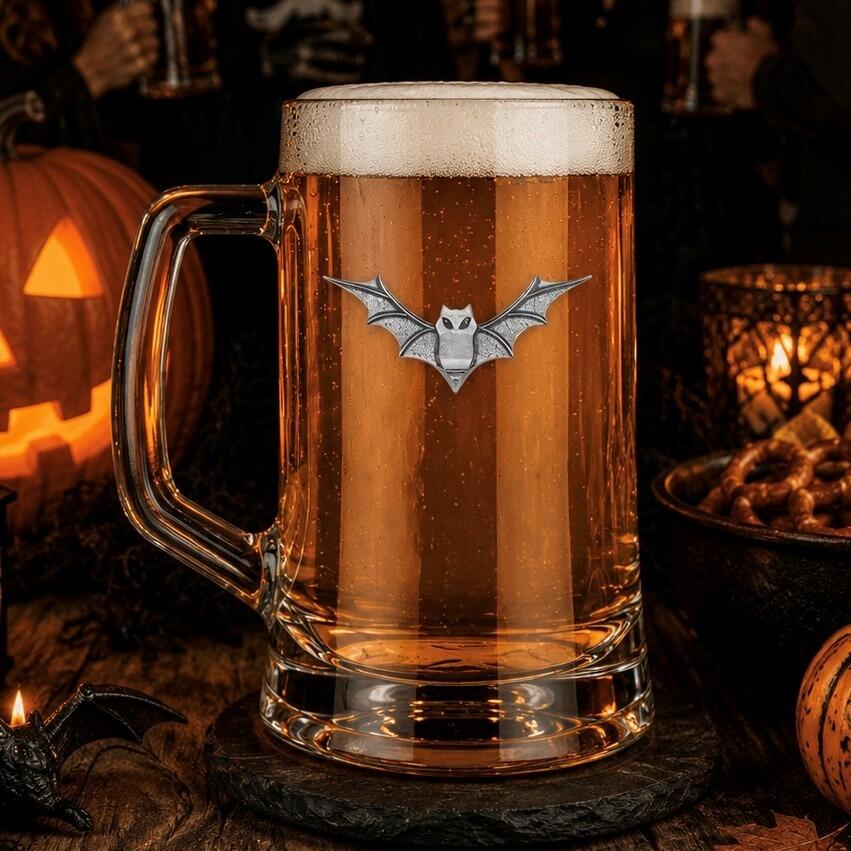 A clear glass beer tankard with a sturdy handle, filled with amber lager and a thick white foam head, featuring a detailed pewter emblem of a bat with outstretched wings