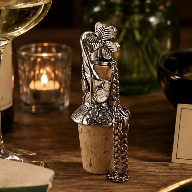 Pewter wine pourer and cork stopper featuring a sculpted traditional Irish shamrock top and engraved clover designs, displayed on a wooden table beside a glass of white wine and a lit candle