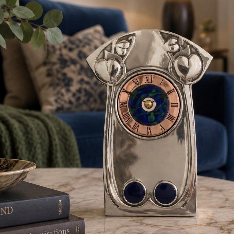An Arts and Crafts style pewter mantel clock designed by Archibald Knox, featuring a polished pewter frame with a widened, decorative top displaying embossed heart-shaped or plant leaf motifs. The clock face has a copper chapter ring with Roman numerals surrounding a swirling blue and green enamelled centre while the base is accented with two inset dark blue enamelled roundels