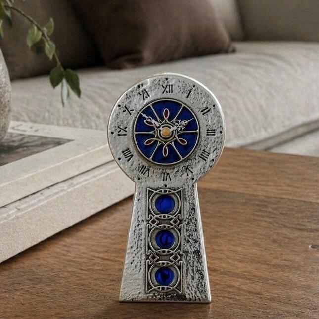 An Arts and Crafts style pewter mantel clock designed by Archibald Knox, featuring a hammered texture and a keyhole-shaped silhouette. The circular clock face displays Roman numerals around a vibrant blue enamelled centre with a gold-toned starburst motif, while the tapered base is vertically decorated with three inlaid blue enamelled roundels within square Celtic knots