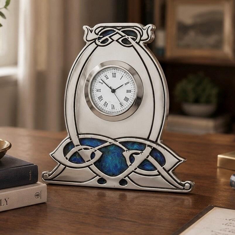 An Arts and Crafts style pewter mantel clock designed by Archibald Knox, featuring a bulbous, horseshoe-shaped frame with an integrated wide base. The clock has a circular white face with black Roman numerals and is accented with intricate Celtic knotwork, including an enamelled top detail and a large, vibrant blue and green enamelled panel stretching across the bottom