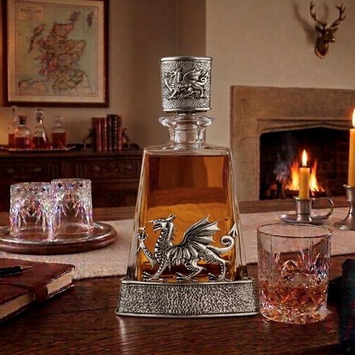 A clear glass spirits decanter filled with amber liquid, sitting in a hammered-effect pewter base. The front of the decanter is adorned with a large, detailed pewter relief of a Welsh dragon with outspread wings. The decanter is topped with a glass stopper encased in a matching textured pewter sleeve featuring an embossed Welsh dragon. On the wooden table beside it sits a cut-glass tumbler containing whisky
