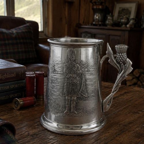 A traditional Scottish pewter tankard featuring a detailed embossed relief of a bagpiper in full Highland dress playing against a mountainous landscape. The tankard is defined by its unique, sculptural handle shaped like a stylized thistle flower with detailed prickly leaves