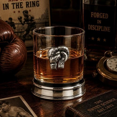 Whisky tumbler with pewter boxing gloves emblem and polished base, partially filled with amber liquid