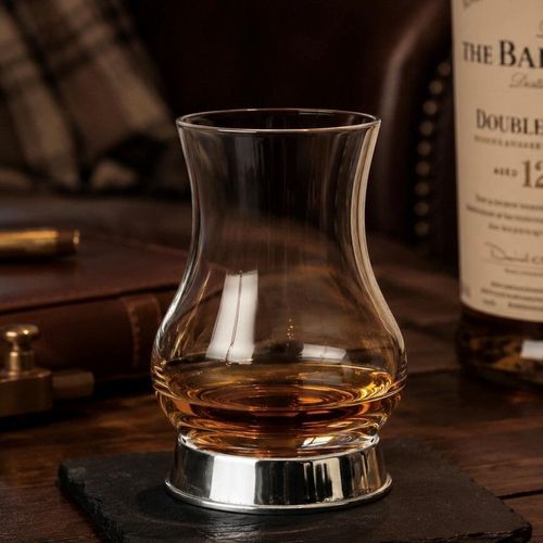 A tulip-shaped whiskey taster glass with an amber pour, resting in a sleek, polished plain pewter base, set against a distillery background