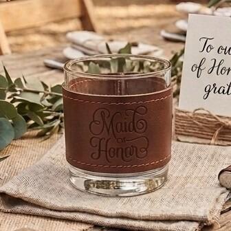 Glass tumbler wrapped in a brown stitched leather sleeve engraved with the words "Maid of Honor" in elegant script