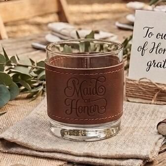 Glass tumbler wrapped in a brown stitched leather sleeve engraved with the words "Maid of Honor" in elegant script